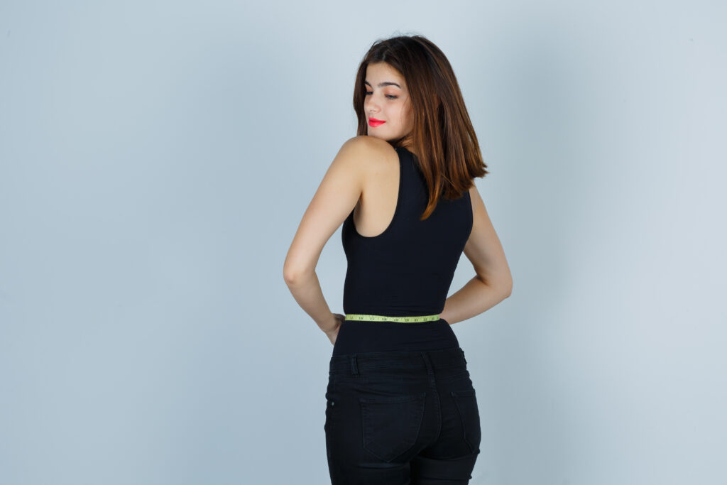 young girl holding measuring tape around her waist, looking over shoulder in black top, pants and looking alluring. front view.